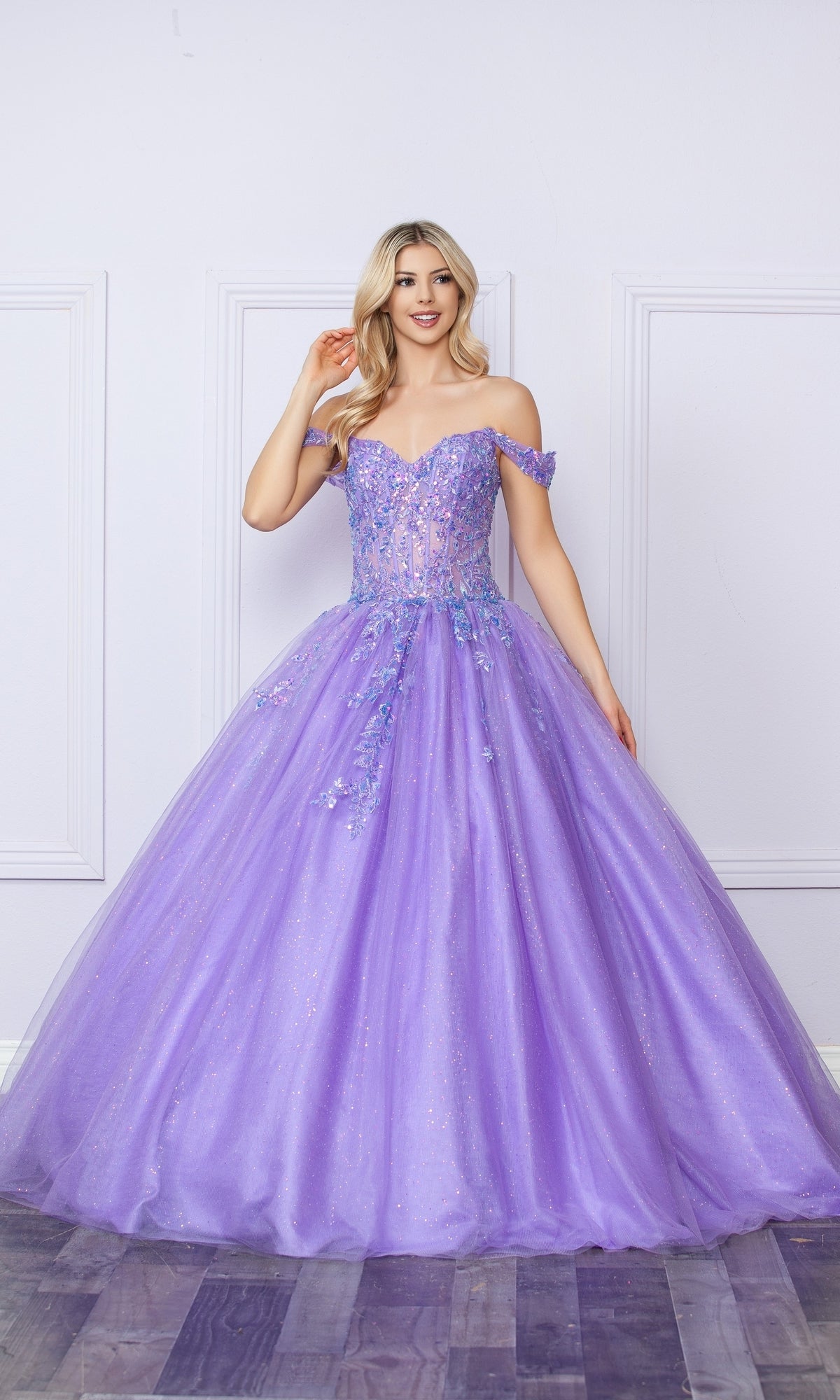 A woman stands indoors in the Bright Off-the-Shoulder Long Prom Ball Gown H1349, a floor-length lavender dress with floral embellishments and a full tulle skirt—perfect for elegant occasions.